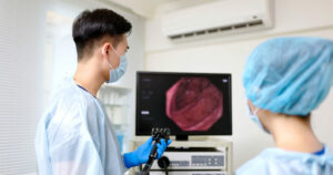 asian male doctor wearing protective gown is doing colon cancer screening and discussing surgical situation with nurse through digital tablet in hospital