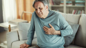An elderly man is experiencing heartburn while sitting on a couch at home.